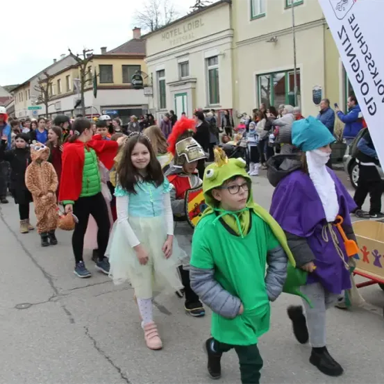Eine Gruppe von Kindern in verschiedenen Kostümen läuft während eines Umzugs die Straße entlang, mit Gebäuden und Schaulustigen im Hintergrund.