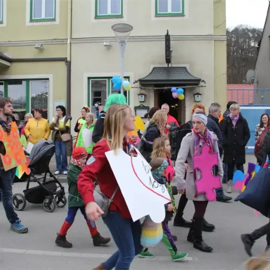 Eine Menschenmenge, einige in Kostümen, geht bei einer Parade auf einer Straße. Im Hintergrund befinden sich Gebäude, Luftballons und ein Kinderwagen.