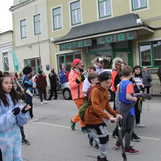 Eine Gruppe von Kindern und Erwachsenen in verschiedenen Kostümen geht vor dem Hotel Loibl die Straße entlang. Einige halten Flaggen und Tennisschläger. Autos sind in der Nähe geparkt.