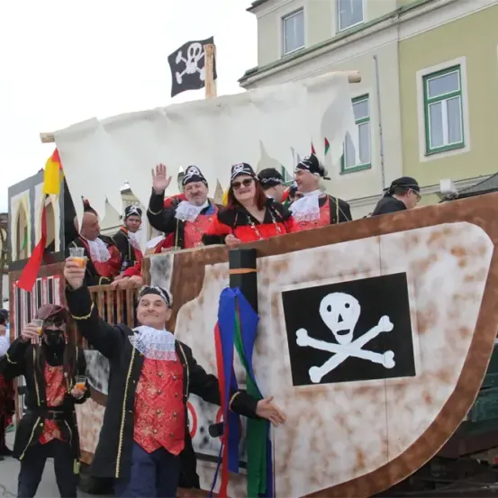 Eine Gruppe von Menschen, die als Piraten verkleidet sind, bei einer Parade, winken und Getränke halten, vor einem Gebäude mit vielen Fenstern.