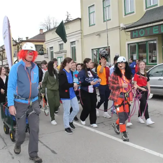 Eine Gruppe von Menschen, einige in Kostümen, geht bei einer Parade die Straße entlang. Eine Person schiebt einen Wagen, eine andere hält eine Seil. Gebäude säumen die Straße, eines ist mit 'Hotel' gekennzeichnet.