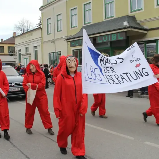Eine Gruppe von Menschen in roten Kostümen mit Masken zieht bei einer Parade. Sie halten ein Banner mit der Aufschrift "Haus der Beratung" und ein Schild mit dem Wort "LBG". Die Gruppe läuft auf einer Straße.