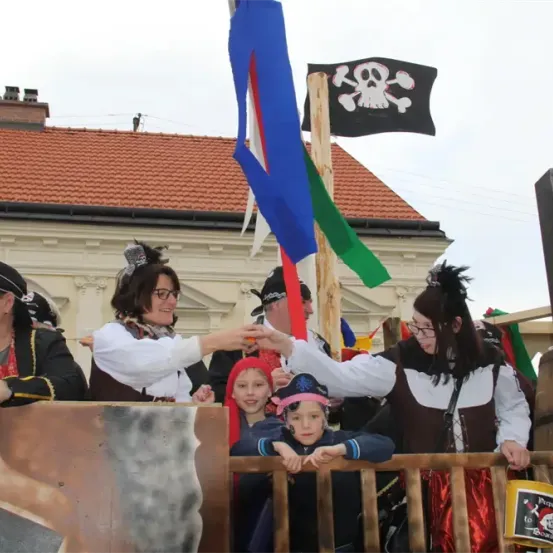 Eine Gruppe von Menschen in Piratenkostümen steht an einer Holzbarrikade, einige halten sich an den Händen. Hinter ihnen weht eine Flagge mit Totenkopf und Knochen. Eine Frau reicht einer jungen Mädchen mit Piratenhut etwas.