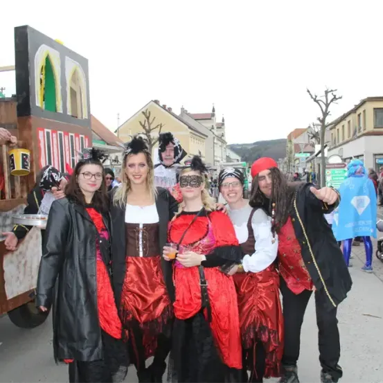 Fünf Menschen in Kostümen stehen auf der Straße, zwei halten Getränke. Dahinter ein Wagen mit Menschen und einem Schild. Gebäude und Bäume sind im Hintergrund.