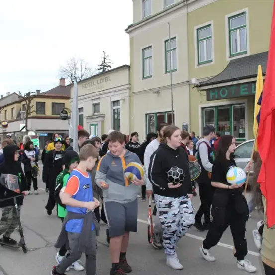 Eine Gruppe von Kindern und Erwachsenen geht auf der Straße. Sie halten Sportausrüstung wie Bälle und Schläger. Gebäude mit dem Wort Hotel befinden sich an der Seite.