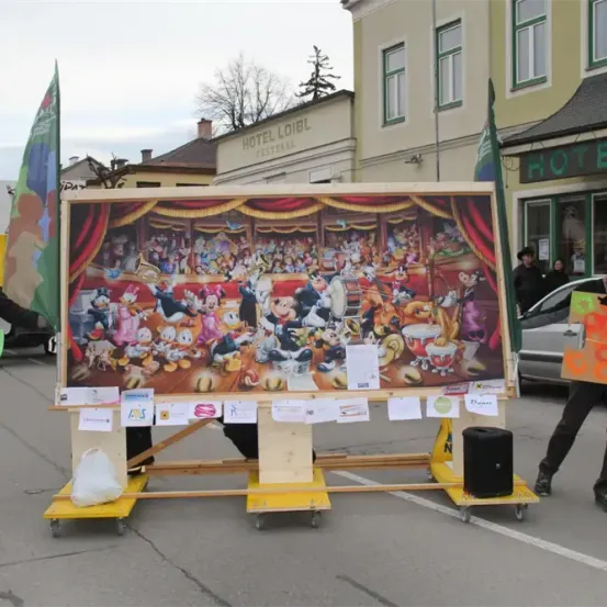 Ein großes Disney-Themengemälde auf einem Holzwagen in einer öffentlichen Straße. Ein Mann hält ein Schild. Dahinter Gebäude mit Fahnen und ein geparktes Auto.