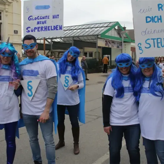 Vier Menschen in Superheldenkostümen stehen auf einer Straße mit Schildern. Zwei Schilder werden von der Gruppe gehalten, eines mit der Aufschrift 'Gloggnitz hat neue Superhelden' und das andere 'Retten Sie mit Ihrer Steuer'.