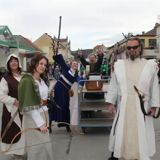 Eine Gruppe von Personen in mittelalterlichen Kostümen, darunter eine Frau mit einem Bogen, steht auf einer Straße. Hinter ihnen sind Menschen in Kostümen und ein Mann mit einem Karren zu sehen.