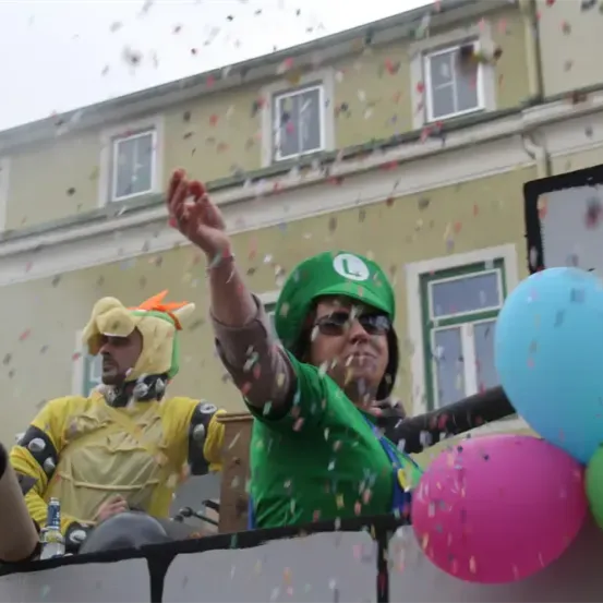 Eine Person in einem grünen Mario-Kostüm feiert im Freien mit Konfetti und Luftballons, neben einer anderen Person in einem gelben Kostüm. Sie stehen auf einer Plattform, mit Gebäuden im Hintergrund.