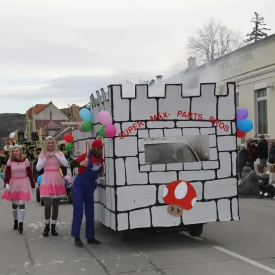 Eine Gruppe von Menschen in Kostümen geht bei einem Umzug mit. Ein wie eine Burg geformter Wagen mit einem Pilzcharakter ist vor ihnen.