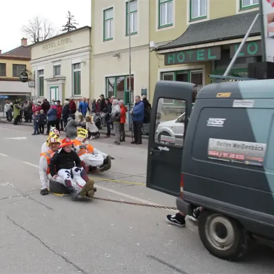 Ein Van zieht eine Gruppe von Menschen, die auf Reifen sitzen, auf einer Straße, mit Zuschauern im Hintergrund. Ein Gebäude mit einem Schild, das Hotel Loibel liest, ist sichtbar.