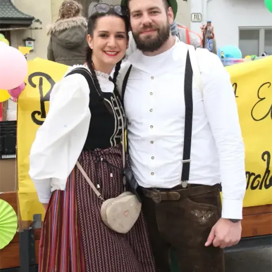 Ein Paar steht eng beieinander und lächelt für die Kamera, gekleidet in traditionelle bayerische Tracht. Die Frau trägt ein langes Kleid mit einem weißen Oberteil und der Mann trägt ein weißes Hemd mit Hosenträgern und braune Hosen. Sie stehen vor einem bunten Hintergrund mit Luftballons und einem gelben Banner.