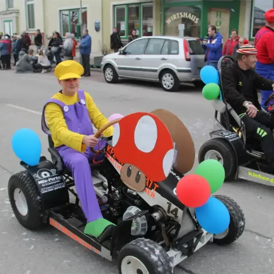 Eine Parade mit zwei Personen auf Go-Karts, einer in Mario-Kostüm, der andere in rotem Overall, beide mit Luftballons. Dahinter steht ein silberner Wagen auf der Straße.