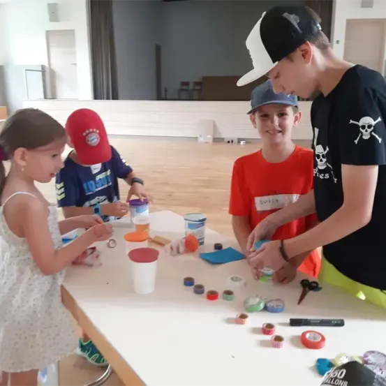 Drei Kinder stehen um einen Tisch mit Bastelmaterial. Ein Mädchen in einem weißen Kleid und zwei Jungen, einer trägt eine rote Kappe und der andere eine schwarze und weiße Kappe.