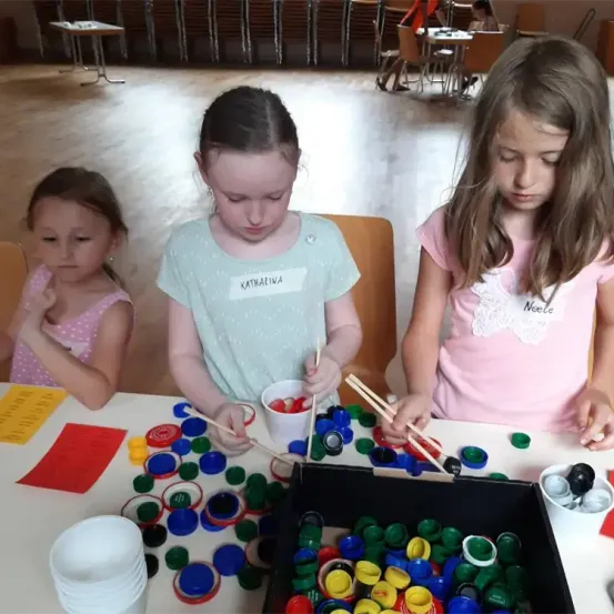 Drei Mädchen sind in ein Spiel mit Flaschenkappen am Tisch verwickelt. Sie benutzen Chopsticks, um die Kappen zu bewegen. Der Tisch ist mit bunten Flaschenkappen und Papieren bedeckt.