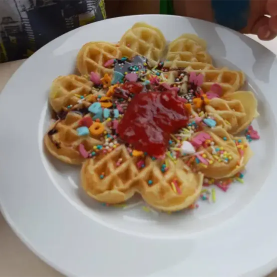 Ein Teller mit Waffeln, die in Form einer Blume angeordnet sind, mit Schokoladensirup, roter Ketchup und bunten Streuseln obenauf.