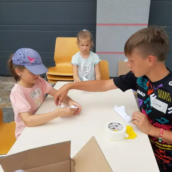 Drei Kinder sitzen an einem Tisch. Ein Junge hält ein Stück Papier und ein Mädchen schaut ihn an. Ein weiteres Mädchen steht hinter ihnen.