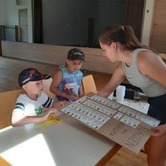 Drei Kinder sind an einer Aktivität beteiligt, wobei ein Mädchen ein Schild mit der Aufschrift Flags of Europe in der Hand hält. Zwei Jungen schauen auf das Schild.