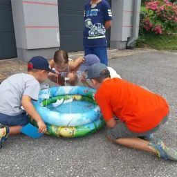 Eine Gruppe von Kindern spielt um einen kleinen aufblasbaren Pool mit Papierbooten in einem Garten mit Blumen und einem Gebäude im Hintergrund.
