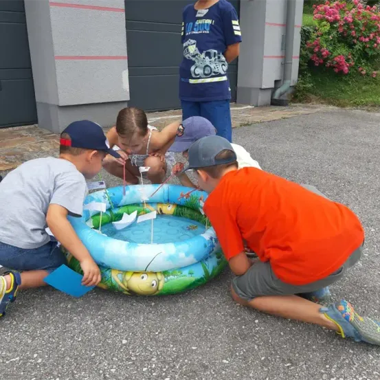Eine Gruppe von Kindern spielt um einen kleinen aufblasbaren Pool mit Papierbooten in einem Garten mit Blumen und einem Gebäude im Hintergrund.