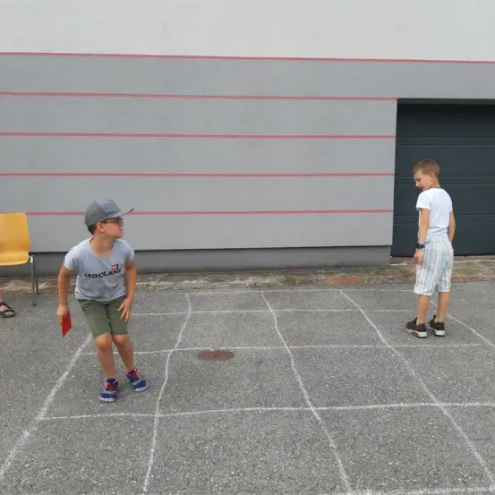 Zwei Jungen spielen Hüpfspiel in einem Parkplatz. Ein Junge trägt ein graues Shirt, grüne Shorts und Turnschuhe. Der andere Junge trägt ein weißes Shirt, Shorts und Turnschuhe. Der Junge auf der linken Seite hält ein rotes Objekt.