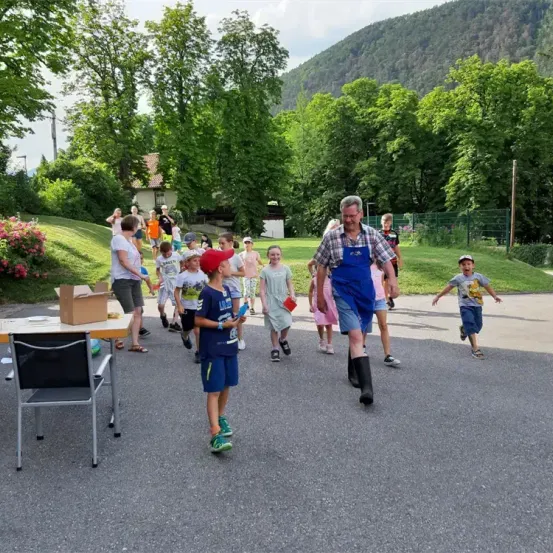Eine Gruppe von Menschen, einschließlich Kindern, läuft in einem Außenbereich. Einige tragen Hüte und Schuhe, und ein Mann in blauer Latzhose geht voraus. Bäume und ein Gebäude sind im Hintergrund zu sehen.
