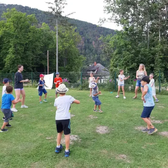 Kinder und Erwachsene spielen auf einer Rasenfläche mit einem Berg und einem Haus im Hintergrund.