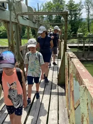 Eine Gruppe von Menschen, darunter Kinder, geht auf einer Holzbrücke über einen Wasserkörper, mit Bäumen im Hintergrund.
