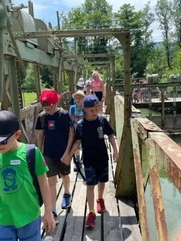 Eine Gruppe von Kindern in Hüten geht Hand in Hand auf einer Holzbrücke und wird von einer Frau und anderen Personen begleitet.