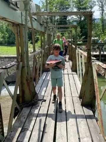 Ein Junge mit Brille geht auf einer hölzernen Brücke und hält ein rotes Buch in der Hand, während andere ihm folgen. Die Brücke ist rostig und hat eine Kette an der Seite. Im Hintergrund sieht man viel Grün.