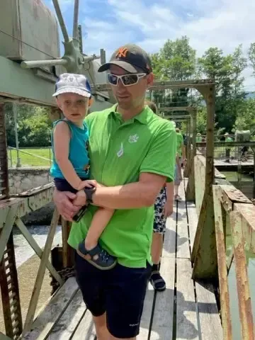 Ein Mann in einem grünen Shirt und mit Sonnenbrille hält ein Kind auf einer Holzbrücke.