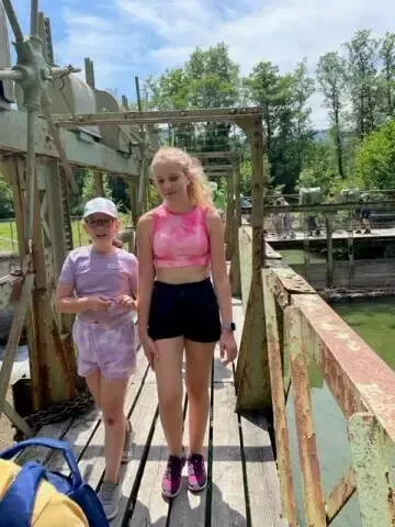 Zwei Mädchen in Sportbekleidung überqueren eine Holzbrücke mit einer rostigen Metallkonstruktion, Bäumen und einem Gewässer im Hintergrund.