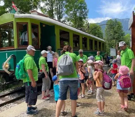 Eine Gruppe von Menschen in grünen Shirts und Hüten steht neben einer Straßenbahn, mit einer Flagge auf dem Zug und einem Berg im Hintergrund.