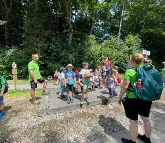 Eine Gruppe von Menschen, einschließlich Kindern, versammelt sich in einem Waldgebiet, wobei einige Rucksäcke tragen. Ein Schild mit weißem Text ist an einem Pfosten montiert.