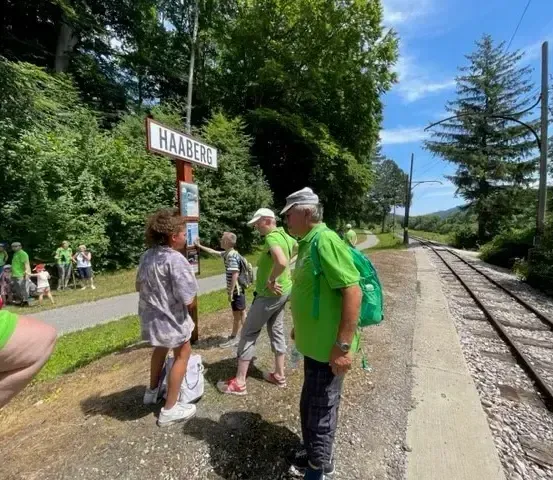 Menschen versammeln sich am Bahnhof Haaberg, an einem sonnigen Tag, mit einem Schild an den Gleisen und Bäumen im Hintergrund.