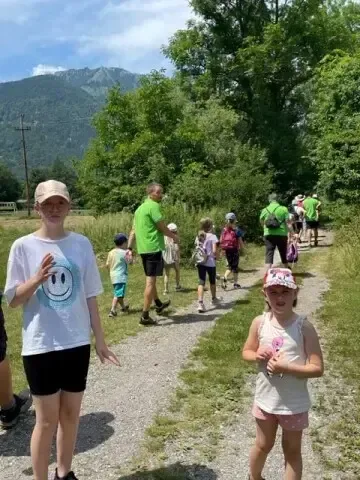 Eine Gruppe von Menschen, einschließlich Kindern, geht auf einem Kiesweg in einem bewaldeten Gebiet mit Bergen im Hintergrund.