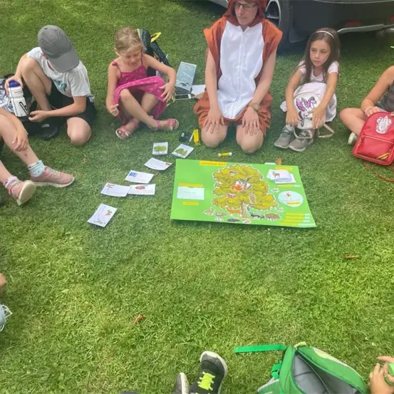 Eine Gruppe von Kindern, darunter eines in einem Fuchskostüm, sitzt auf dem Rasen mit einem Lernspielbrett vor ihnen. Sie haben Rucksäcke und Spielzeug um sich herum verstreut.