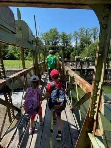 Zwei Kinder mit Rucksäcken überqueren eine Holzbrücke über einen Fluss. Dahinter folgt ein Mann in einem grünen Shirt und einem roten Hut. Bäume und eine Wiese sind im Hintergrund.