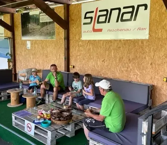 Eine Gruppe von Menschen sitzt auf einer Couch in einem Raum. Ein Mann in einem weißen Hut sitzt auf der Couch und schaut sich das Essen auf dem Tisch an.