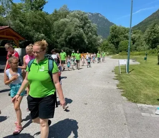 Eine Gruppe von Menschen, einige in grünen Shirts und mit Rucksäcken, geht auf einem gepflasterten Weg. Eine Person fegt den Bürgersteig. Bäume und Berge sind im Hintergrund.