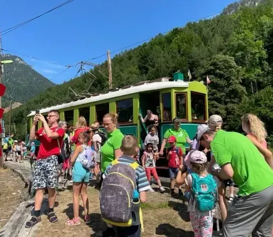 Eine Gruppe von Menschen, einschließlich Kinder mit Rucksäcken, steigen an einem sonnigen Tag in einen grünen Tram ein. Sie sind von üppiger Grünfläche und Bergen umgeben.