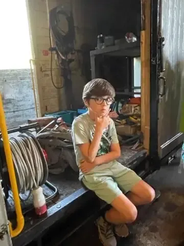 Ein Junge mit Brille sitzt auf der Kante eines Lastwagens und grübelt. Der Lastwagen ist mit verschiedenen Werkzeugen und Gegenständen gefüllt.