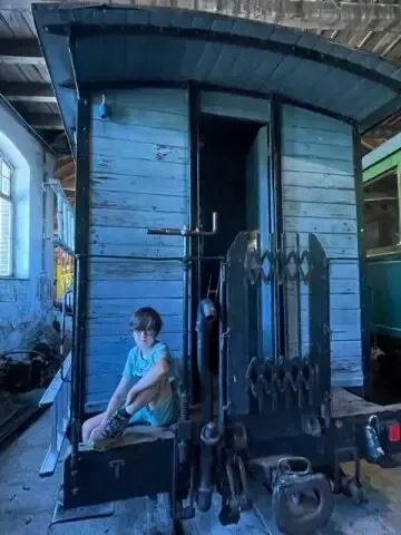 Ein Kind sitzt in einem blauen Zugwagen mit offenen Türen in einer Werkstatt mit einem grünen Zugwagen dahinter.