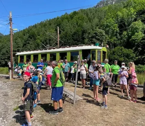 Eine Gruppe von Menschen in grünen Shirts versammelt sich auf einem Schotterplatz vor einem grünen Zug, mit Bäumen und Bergen im Hintergrund.