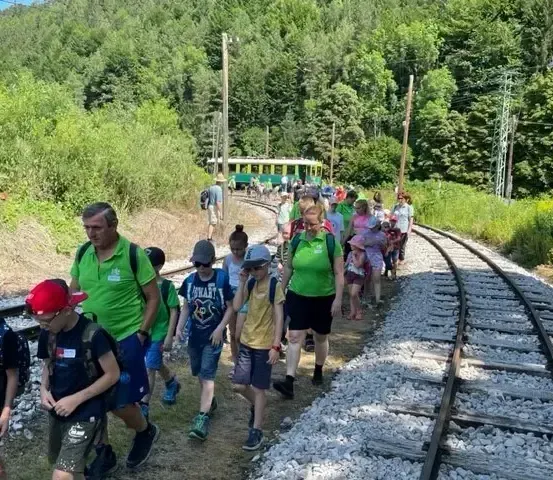 Eine Gruppe von Menschen, einschließlich Kindern, geht entlang einer Eisenbahnschiene. Einige tragen Rucksäcke und Hüte, und ein Zug ist in der Ferne zu sehen.