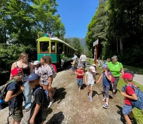 Eine Gruppe von Kindern und Erwachsenen geht auf einem Schotterweg in der Nähe eines Zuges, mit Bäumen und einem klaren blauen Himmel im Hintergrund.