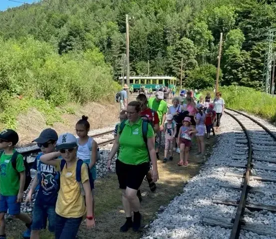 Eine Gruppe von Menschen geht auf einem Pfad in der Nähe einer Bahnstrecke. Die Strecke ist von Grün umgeben. Einige tragen Rucksäcke.