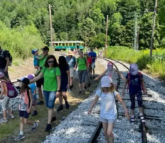 Eine Gruppe von Menschen, einschließlich Kindern, geht auf einem mit Kies bedeckten Bahngleis, mit einem grünen Zug im Hintergrund.