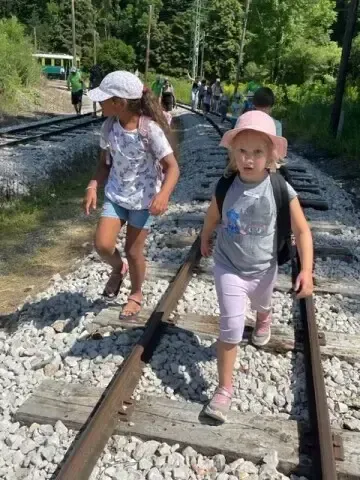 Zwei junge Mädchen gehen auf einem Eisenbahngleis. Sie tragen Hüte und haben Rucksäcke. Im Hintergrund geht eine Gruppe von Menschen entlang der Gleise. Bäume und Gras säumen die Seiten.