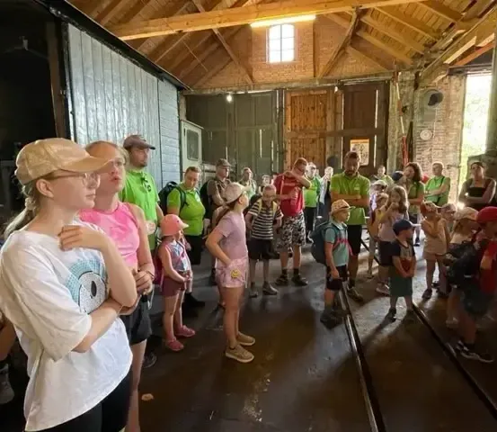 Eine Gruppe von Menschen, wahrscheinlich Touristen, steht und schaut sich etwas in einem Gebäude an. Einige tragen Hüte, Brillen und Turnschuhe. Es gibt Backsteinmauern, Holzbalken und eine offene Tür. Einige Leute tragen grüne Uniformen.